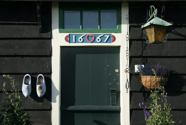 Dark green wooden door with a pair of wooden clogs hanging on the left and a hanging basket filled with lavender hanging on the right