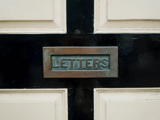 aged brass letterbox that reads letters on it, against a black and white door