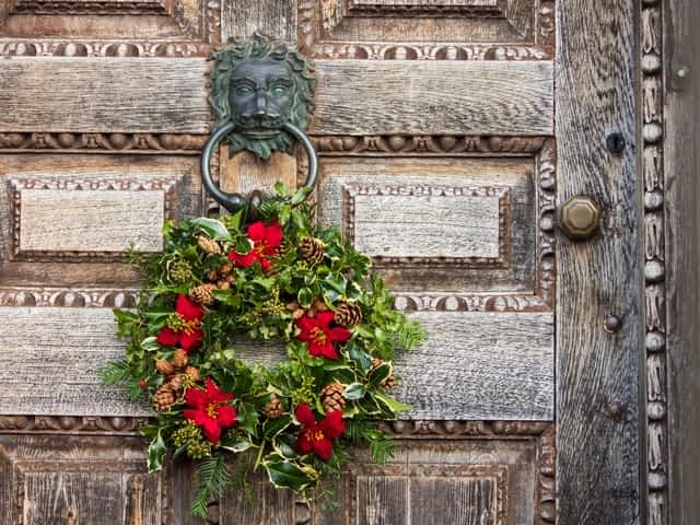 christmas wreath, made of ivy, holly, pinecones and red ribbon against a large distressed oak door with several panels
