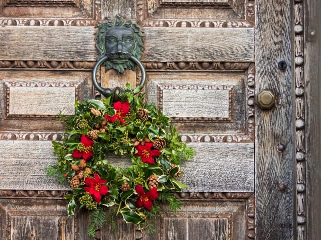 christmas wreath, made of ivy, holly, pinecones and red ribbon against a large distressed oak door with several panels