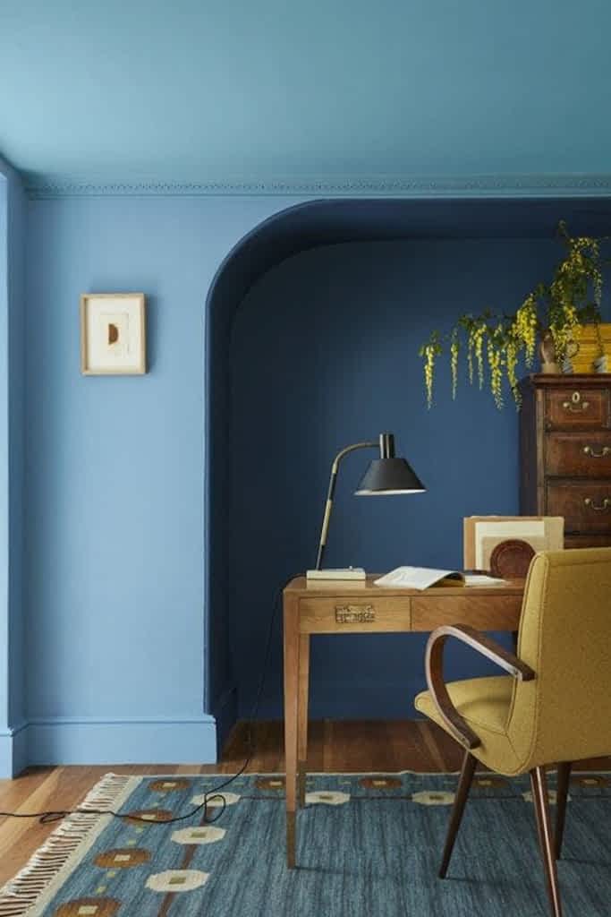 A deep blue painted wall featuring an alcove with a desk, chair and lamp