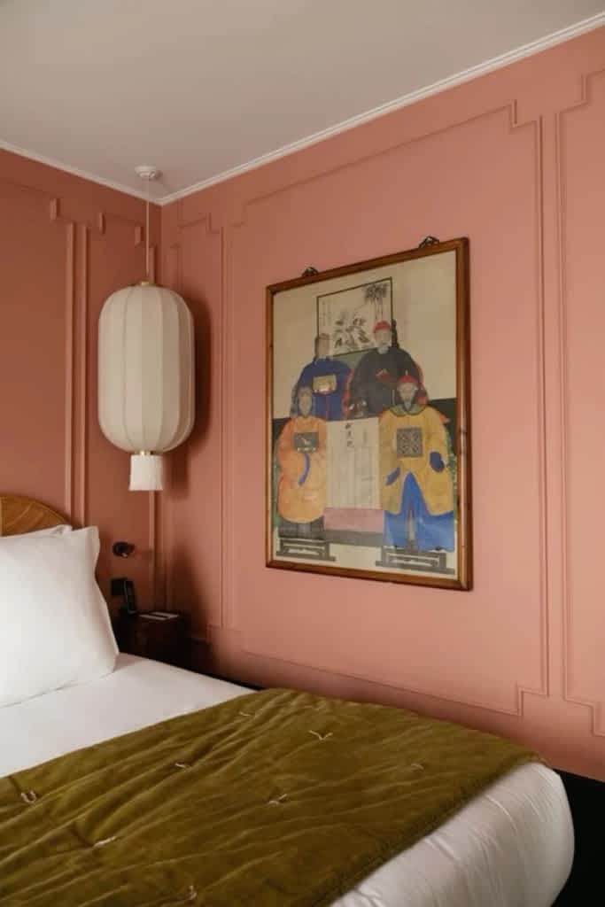 The corner of a pink bedroom showing a large painting on the wall, a large lampshade and the corner of a bed