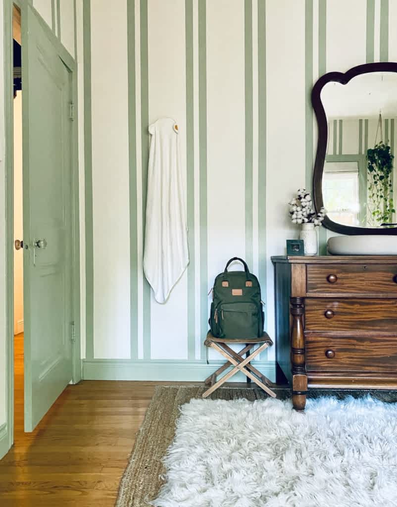 A bedroom with green and white wallpaper, a green door and a chest of drawers and mirror