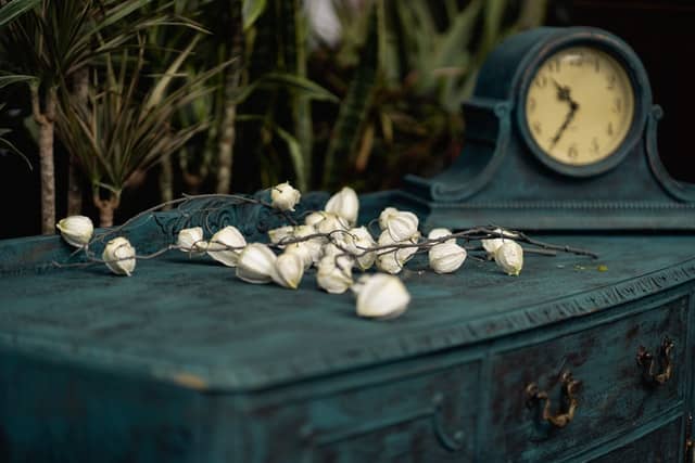 navy dresser with a navy clock atop, adorned with dying white flowers and dracaena plants peeking from behind it